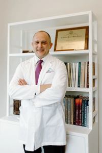Physician in a white lab coat standing with arms crossed in front of a bookshelf with diplomas and medical books.
