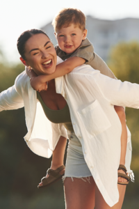 Woman giving a piggyback ride to a smiling young boy outdoors on a sunny day.