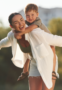 Woman gives a piggyback ride to a smiling child outdoors, sunlight behind them.