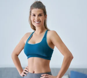 Smiling woman in a teal sports bra and gray leggings with hands on hips in a bright room.