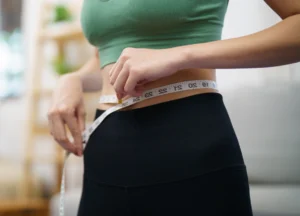 Person measuring their waist with a tape measure while wearing a green sports top and black leggings in a bright room
