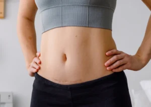 Abdomen with a horizontal surgical scar along the lower belly, wearing a gray sports bra and black pants, hands resting on hips