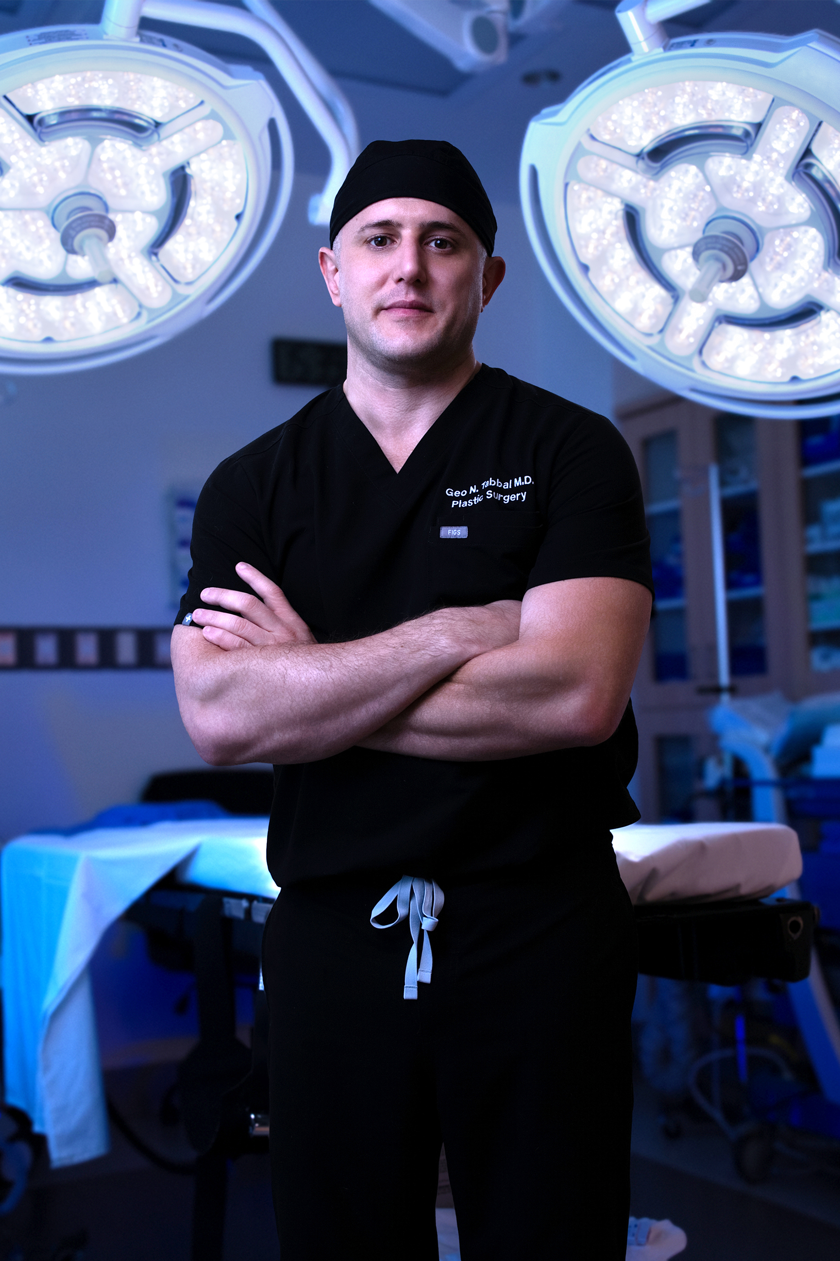 Surgeon in black scrubs and cap stands with arms crossed in an operating room, large surgical lights overhead.