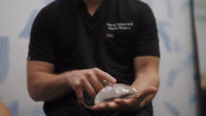 Surgeon in black scrubs holding a silicone breast implant during a medical demonstration.