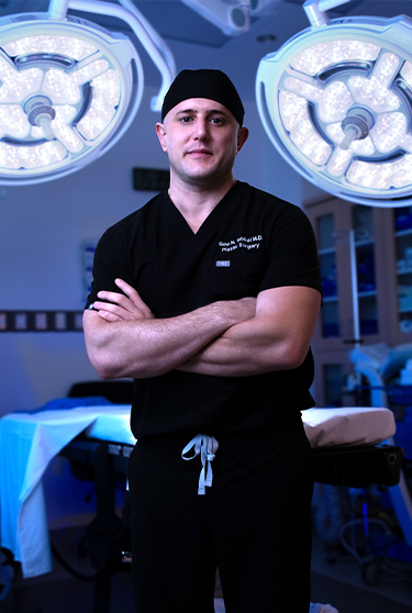 Surgeon in black scrubs and cap stands with arms crossed in an operating room under bright overhead lights.