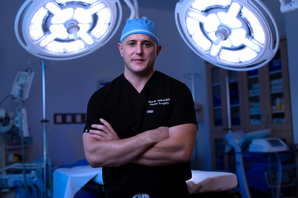 Male surgeon in black scrubs and a blue surgical cap stands with arms crossed under bright overhead surgical lights in an operating room.